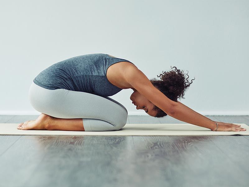 A person in a gray top and leggings performs child's pose, resting on a yoga mat in a minimalist room.