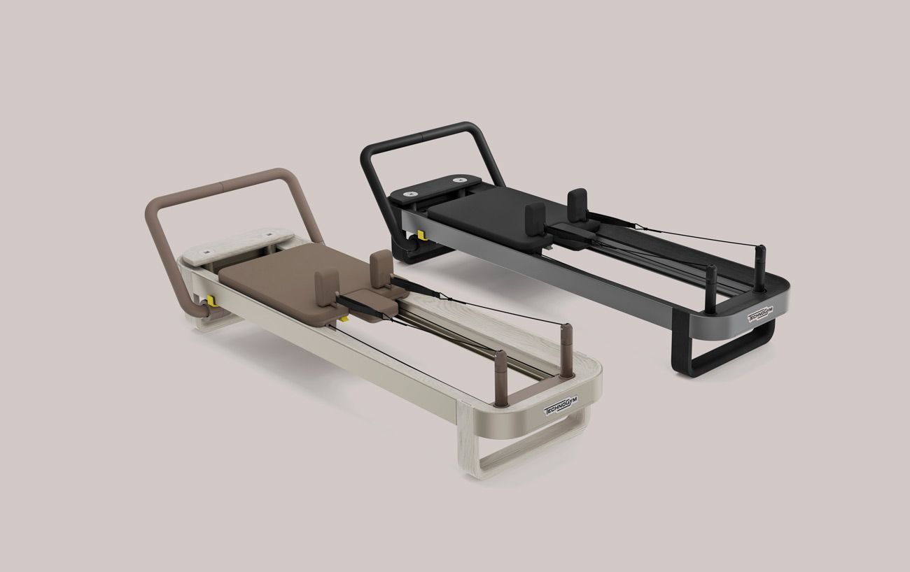 Two Pilates reformers displayed side by side: one in light beige with a brown pad, and one in dark grey with a black pad.