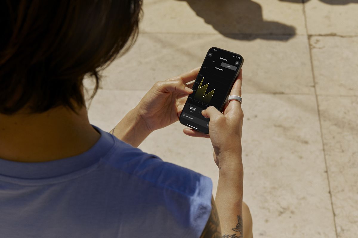 A person in a light blue shirt outdoors holds a smartphone displaying a yellow line graph on a dark background.