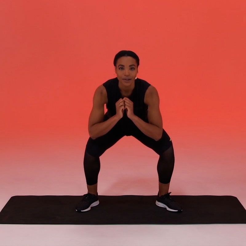 A person in dark workout clothes performs a squat on a black exercise mat against a solid red background.