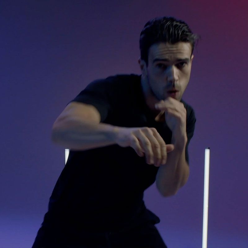 A person in a black t-shirt in a boxing stance against a purple background with two vertical neon lights.