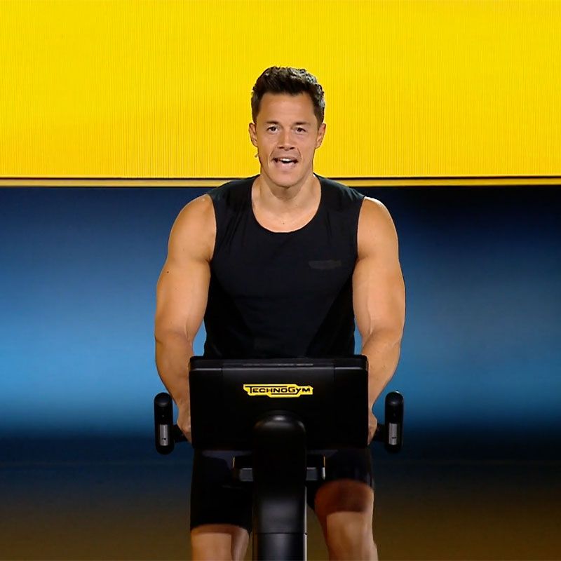 A person in a black tank top exercising on a Technogym stationary bike against a yellow and blue background.