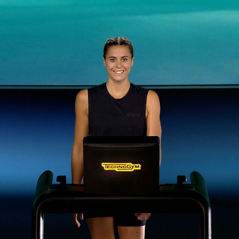A person smiling while standing behind a Technogym exercise machine against a blue background.