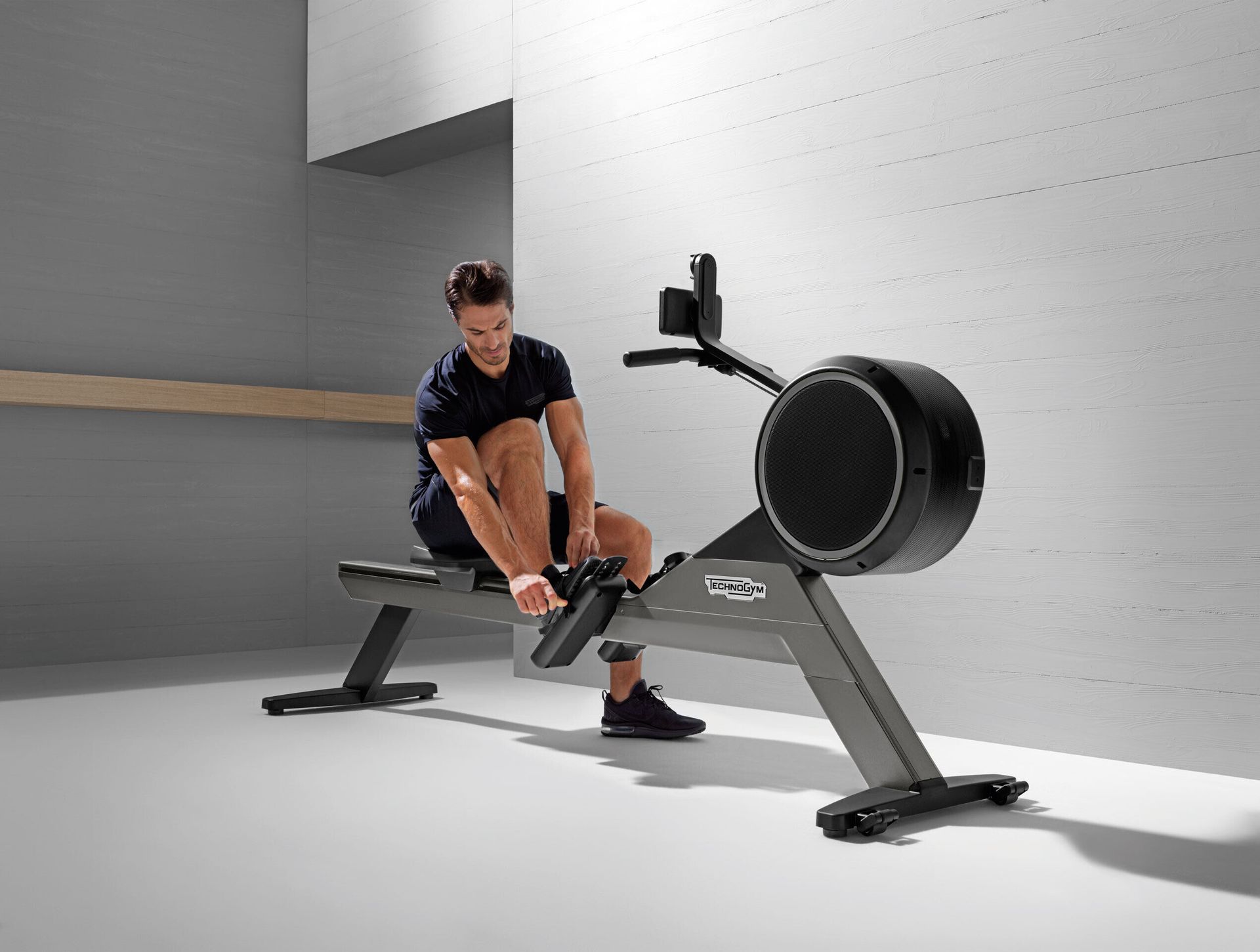 A person in athletic gear sits on a black rowing machine in a minimalist room, adjusting the foot straps.