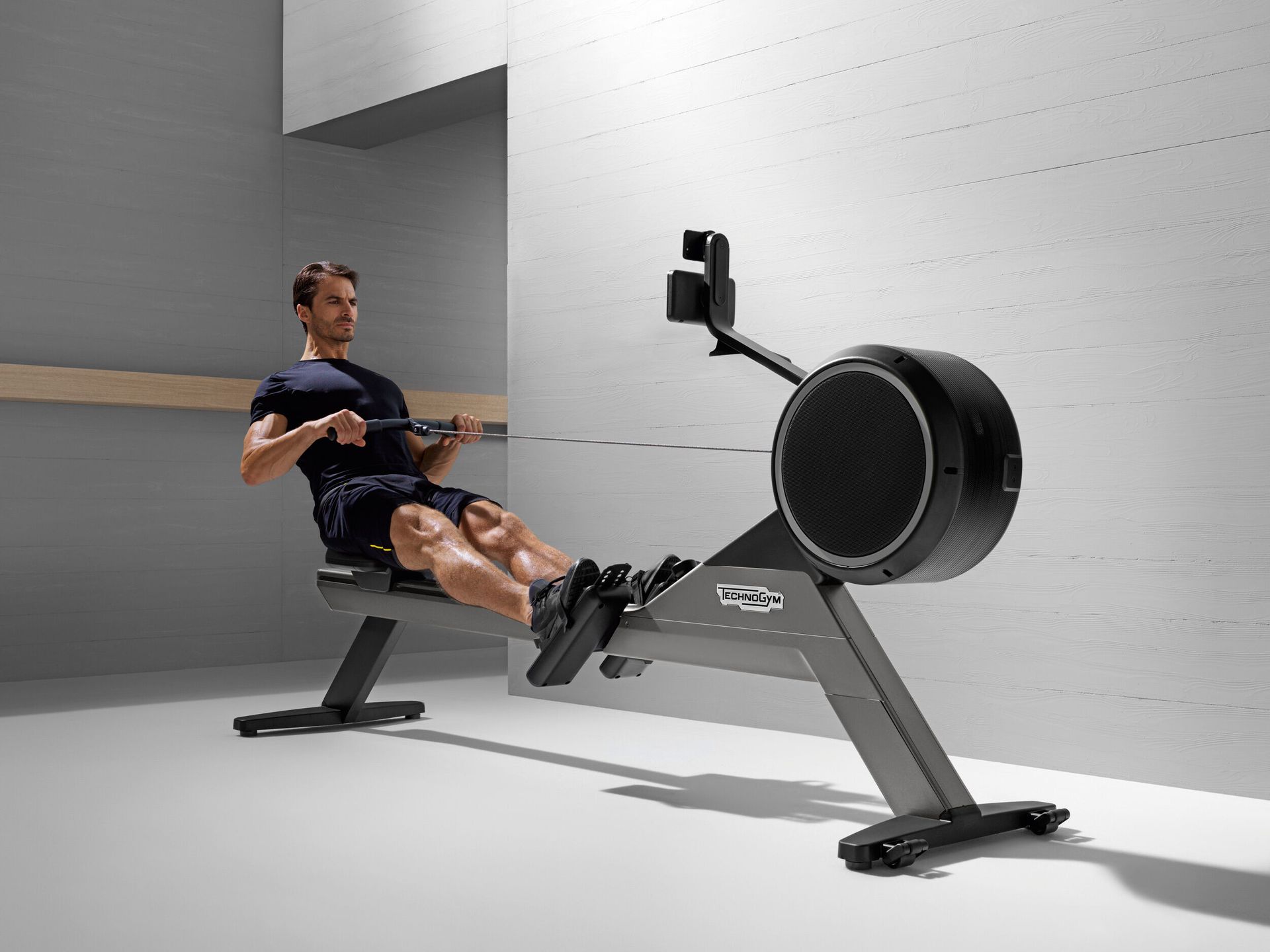 A person using a dark gray rowing machine in a minimalist, light-colored room.