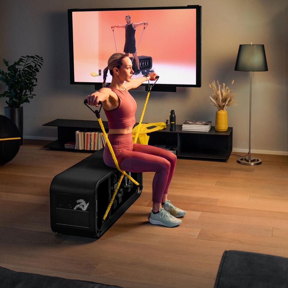A person in workout clothes exercises on a black bench with yellow resistance bands in a living room with a TV display.