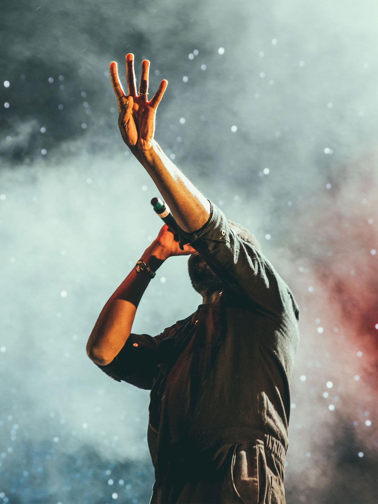 A performer on stage holding a microphone with one hand raised, silhouetted against a smoky background with bright lights.