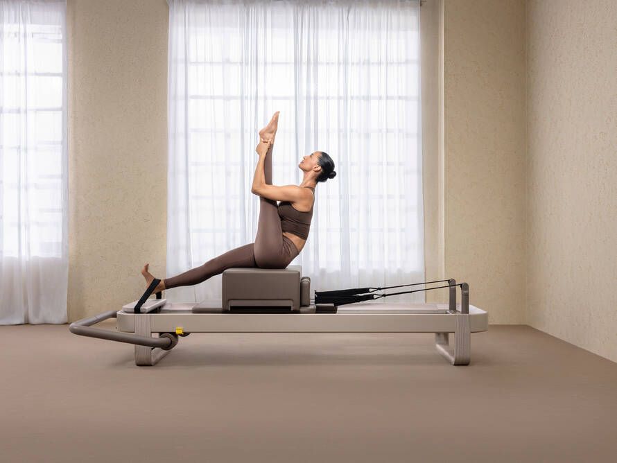 A person performs a Pilates exercise on a reformer machine, sitting on a box with one leg extended vertically.