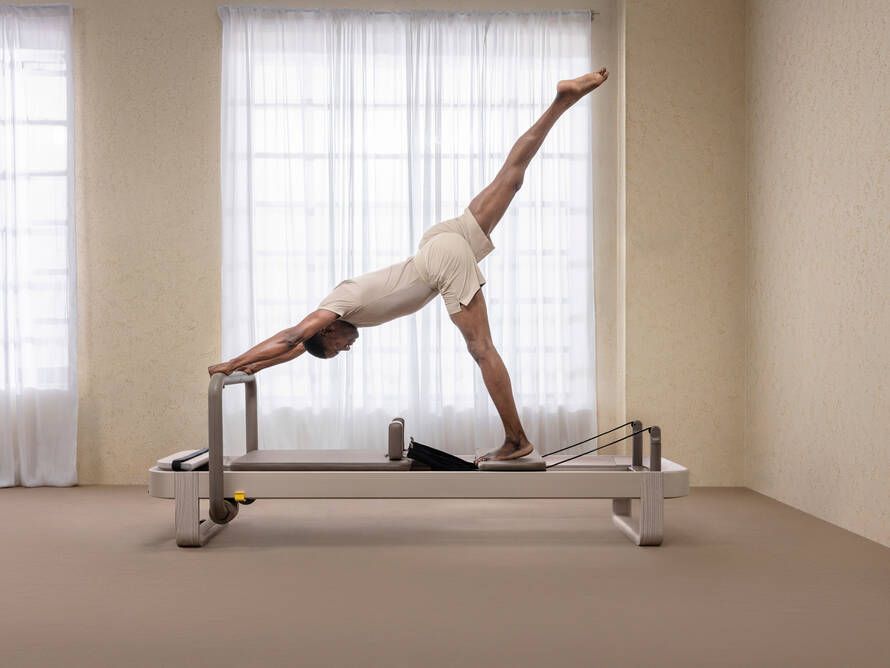 A person in light-colored activewear performs a single-leg pilates exercise on a reformer in a bright, minimalist studio.