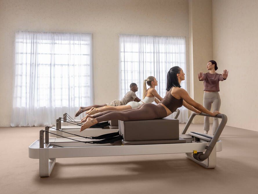 Three people on Pilates reformers performing an extension exercise while an instructor leads the class in a bright studio.