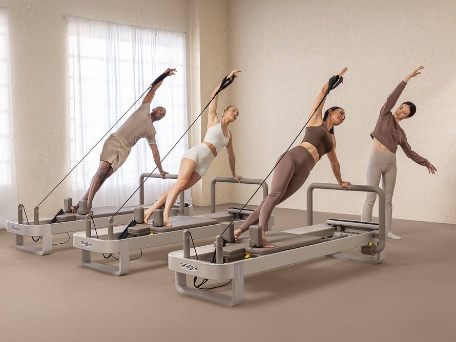 Four people in neutral-toned activewear perform side plank variations on Pilates reformers in a bright, minimalist studio.