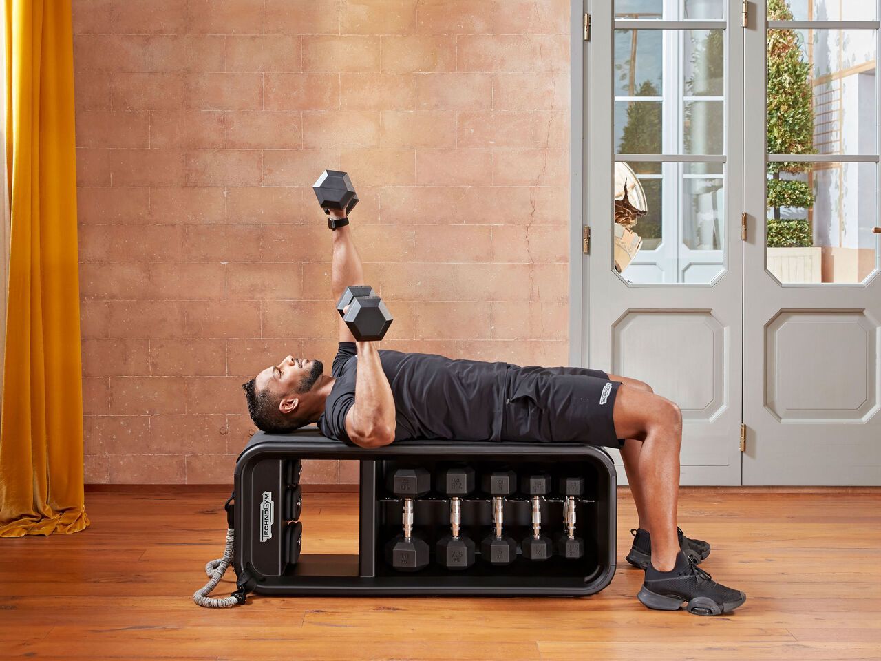 A person lies on a Technogym exercise bench, performing a one-arm dumbbell chest press in a bright, modern room.