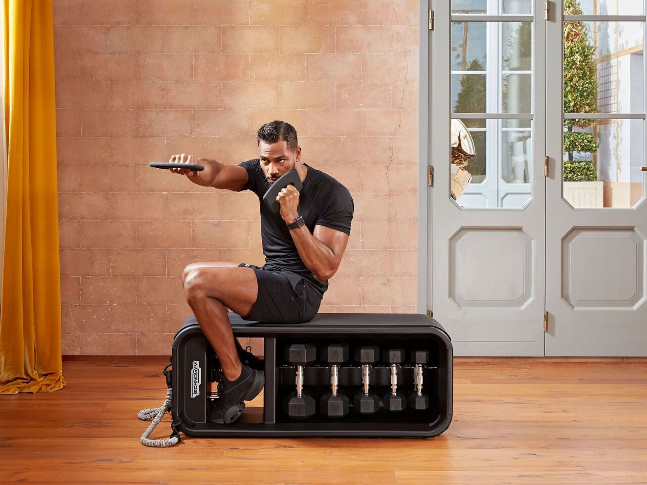 A person sitting on a dark workout bench with dumbbells, extending one arm forward while holding a weight plate.