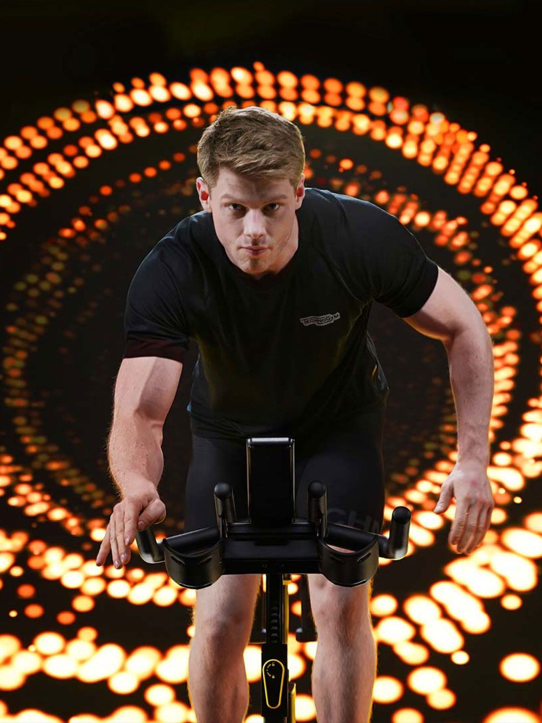 A person riding a stationary cycle in front of a dark background with circular, glowing orange light patterns.