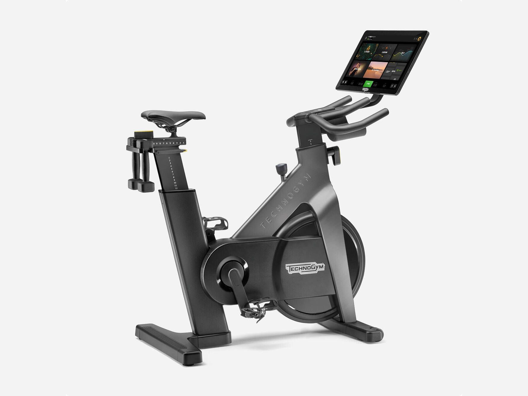 Black Technogym stationary bike with a tablet screen, water bottle holders, and dumbbells against a white background.