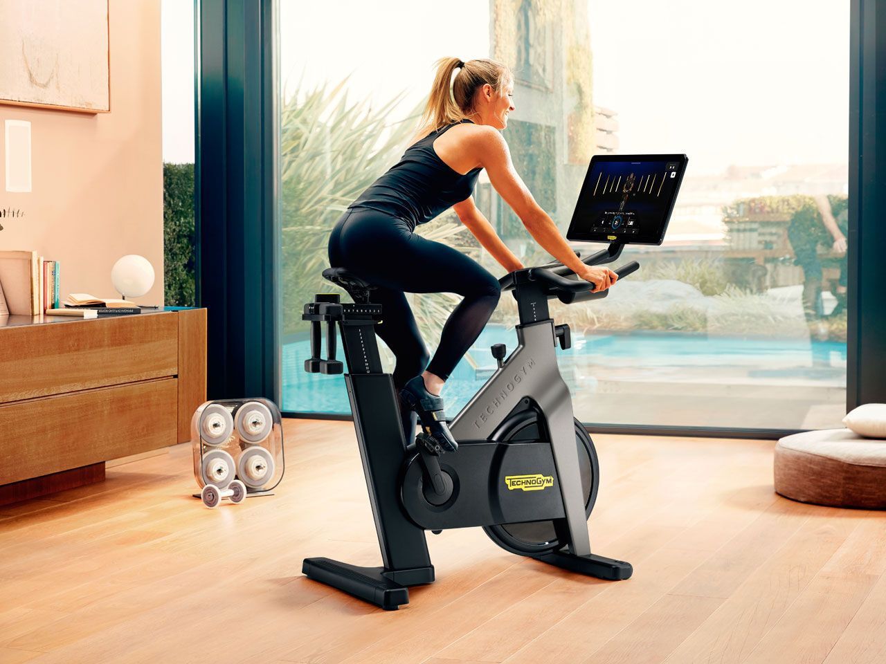 A person exercises on a Technogym stationary bike in a bright, modern room overlooking a pool.