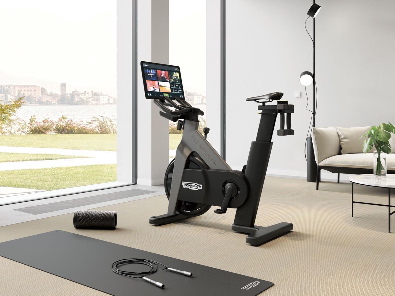 A Technogym indoor exercise bike with a display screen, exercise mat, foam roller, and jump rope in a bright, modern room.