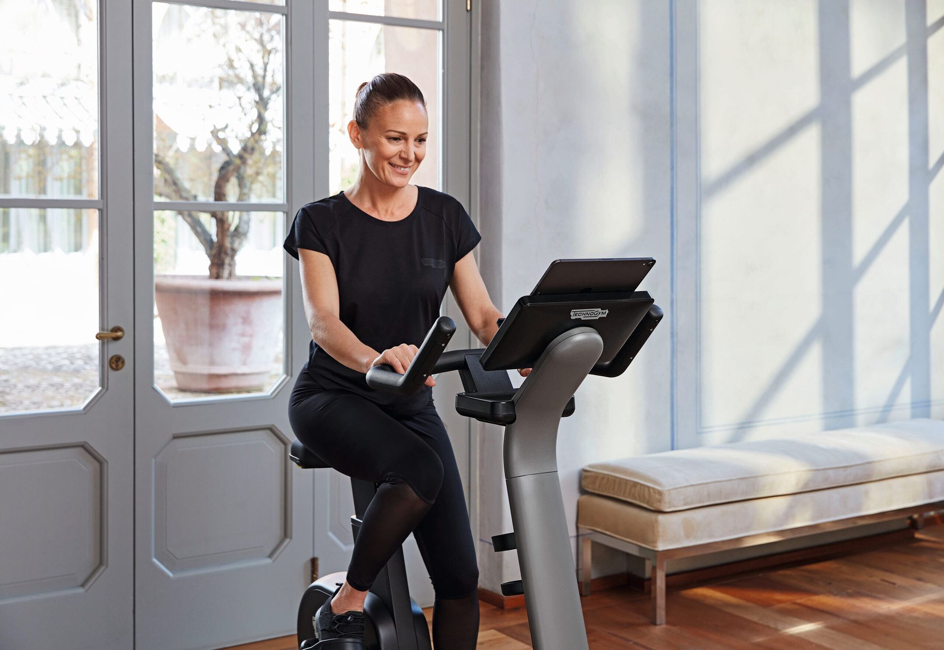 A smiling person exercising on a stationary bike in a bright room with French doors and a bench.