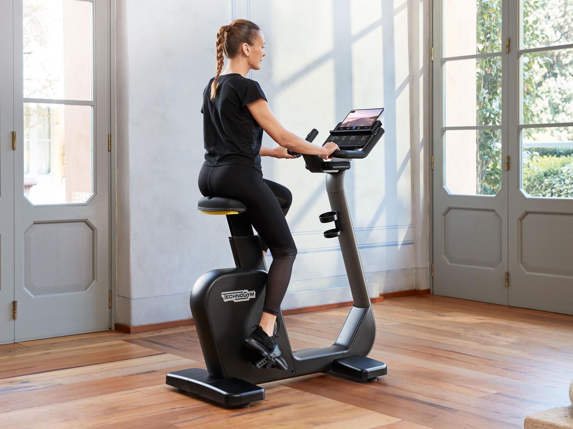 A person sits on a black exercise bike in a bright, modern room with large glass doors and wooden floors.