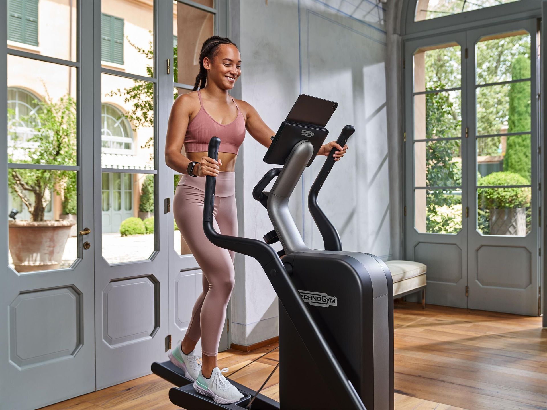 A person in pink athletic wear exercising on a Technogym elliptical machine in a bright room with large glass doors.