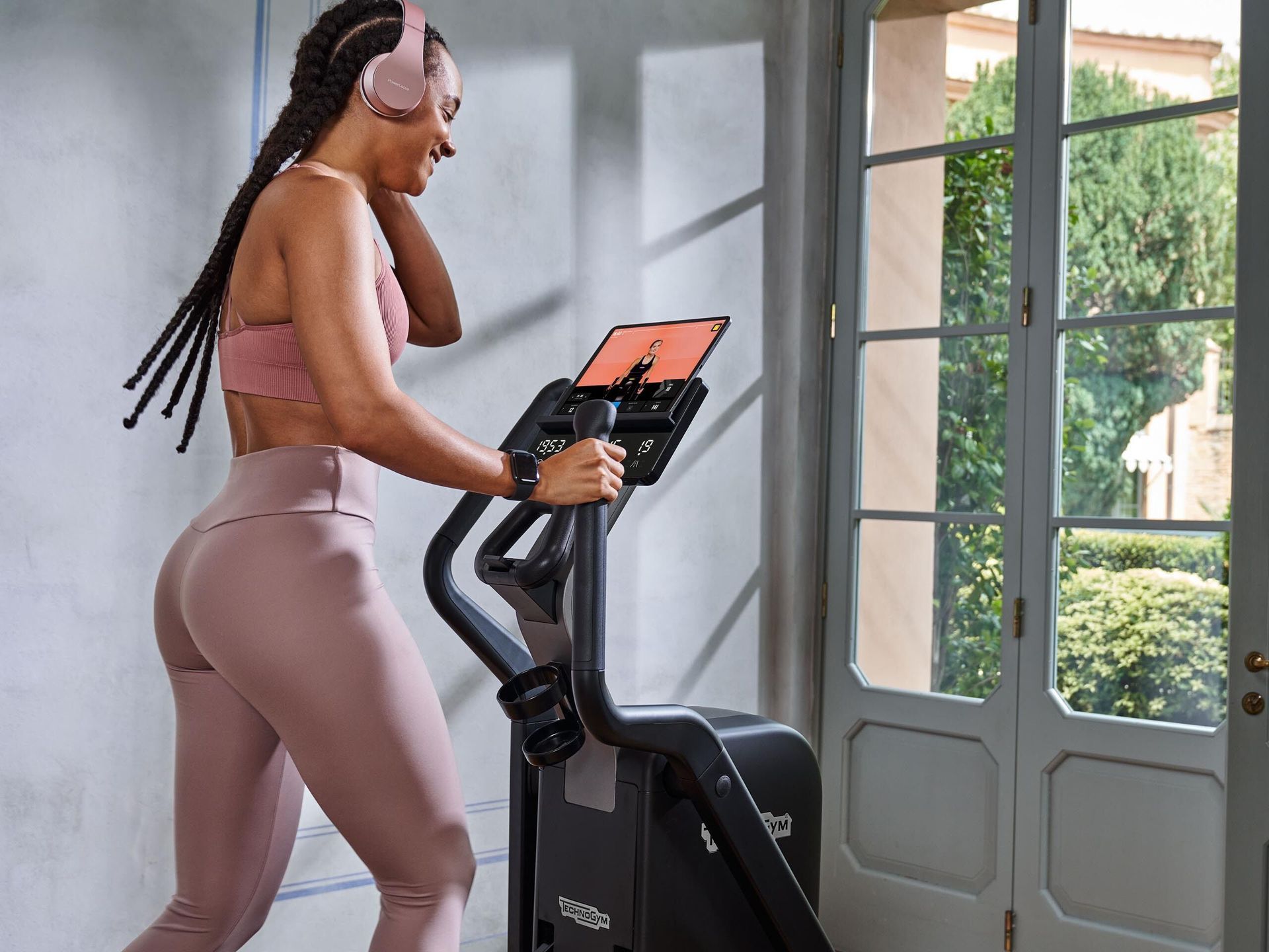 A person in pink athletic wear uses an elliptical machine while wearing headphones, near a doorway with a garden view.