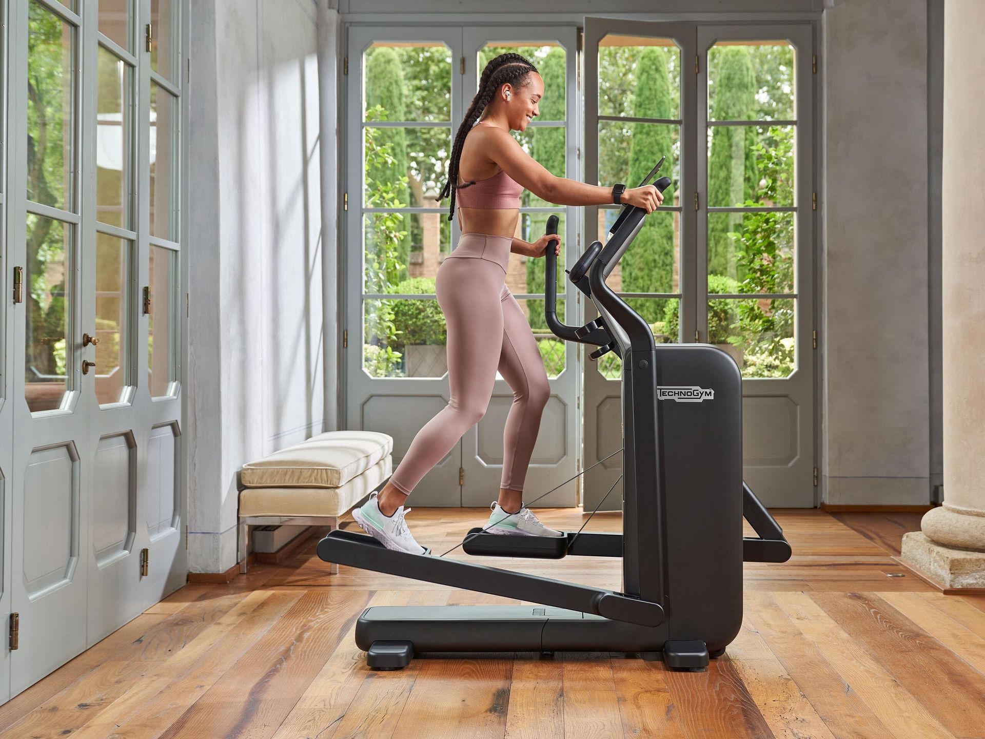 A person in athletic wear exercises on a Technogym elliptical machine in a bright, sunlit room with wooden floors.