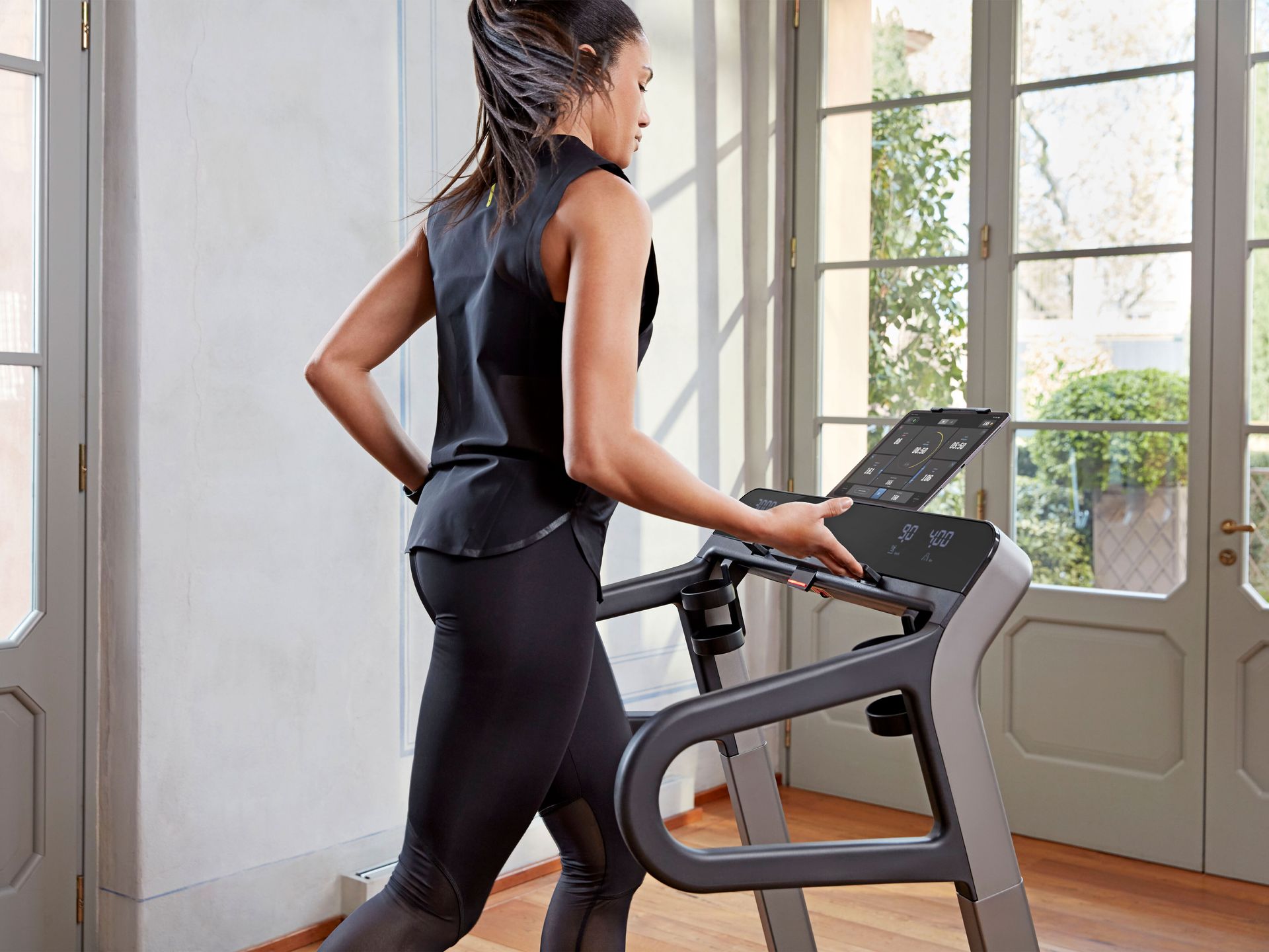 A person in athletic wear exercises on a treadmill in a sunlit room with large glass doors.