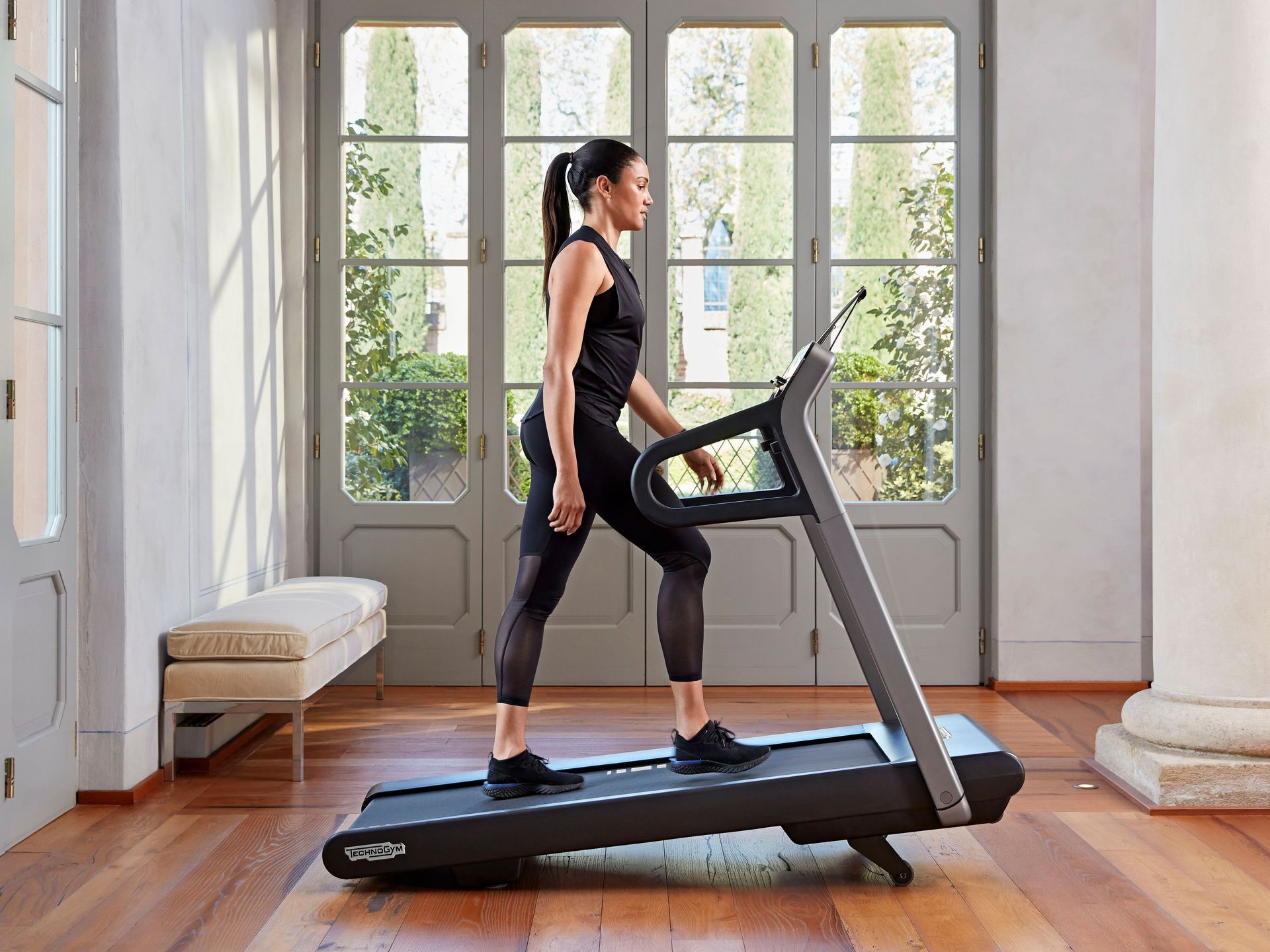 A person in athletic gear stands on a treadmill in a sunlit room with large windows and hardwood floors.