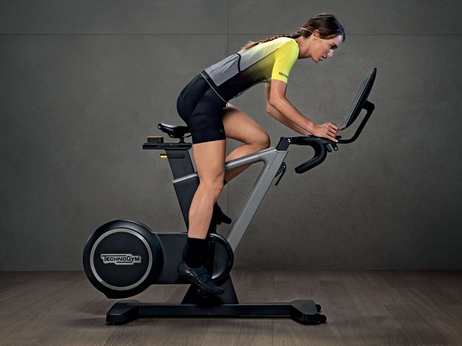 A person in athletic gear riding a Technogym stationary bike against a plain grey background.