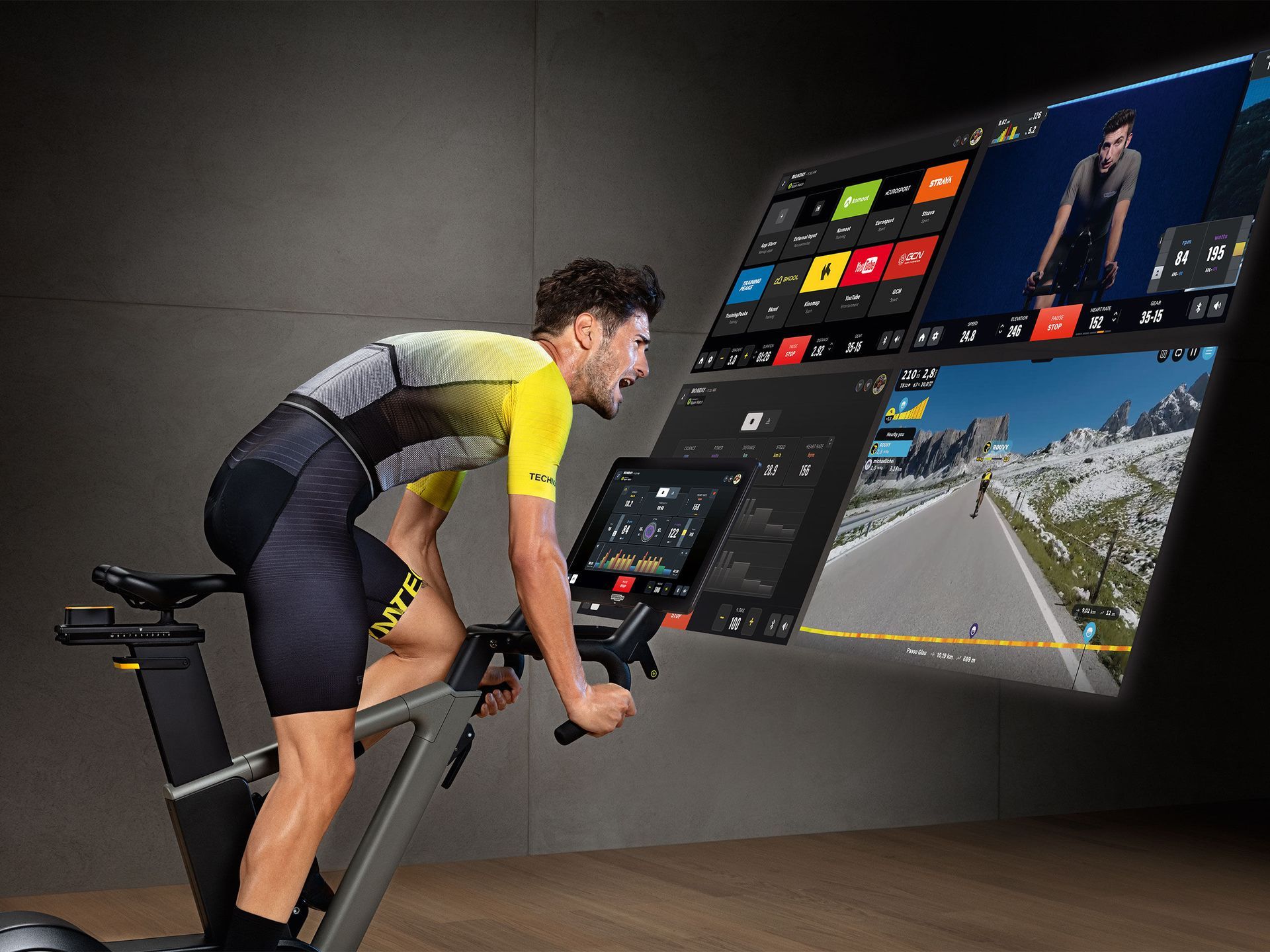 A cyclist in a yellow and black kit trains on an indoor bike while facing a large digital screen showing workout data.