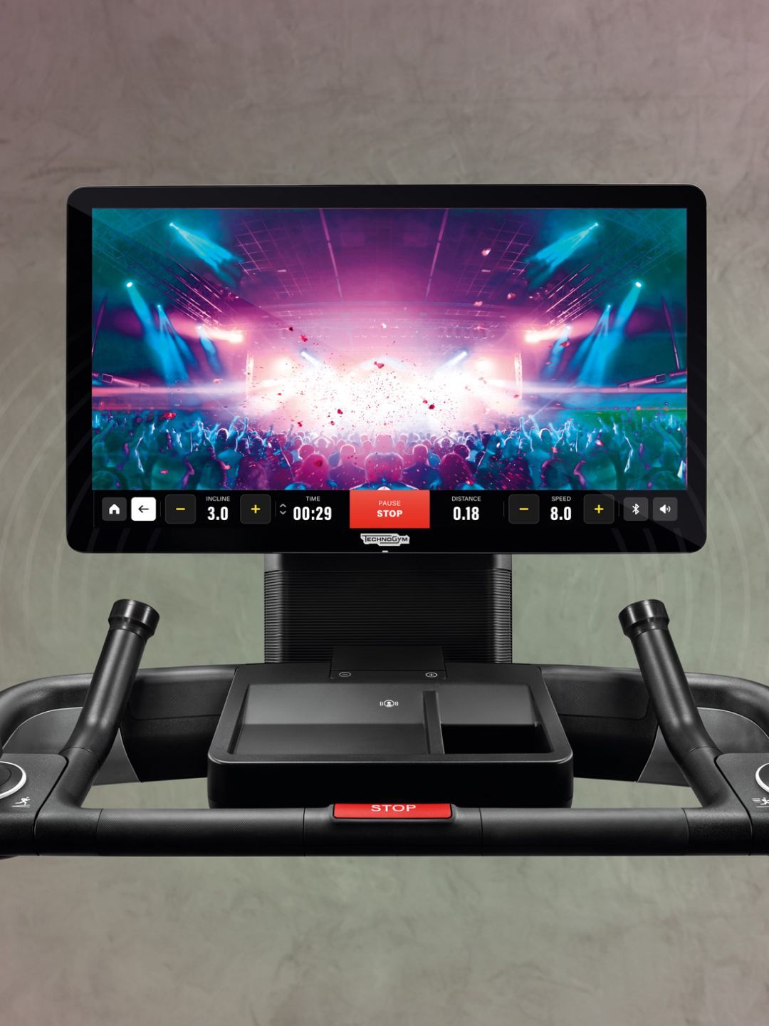 A Technogym treadmill console displays a vibrant concert scene on its screen, set against a neutral, soft grey background.