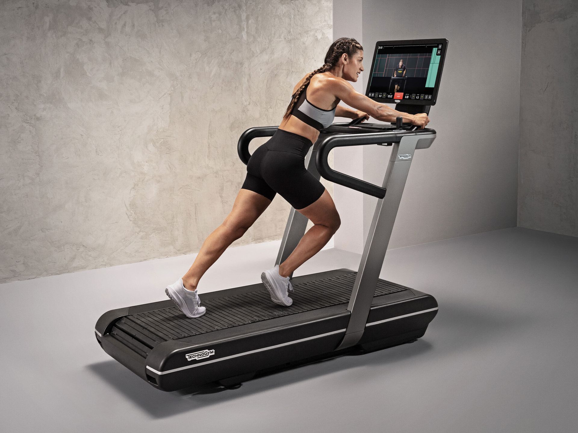 A person exercising on a black Technogym treadmill with an integrated screen, set against a plain, light-colored background.
