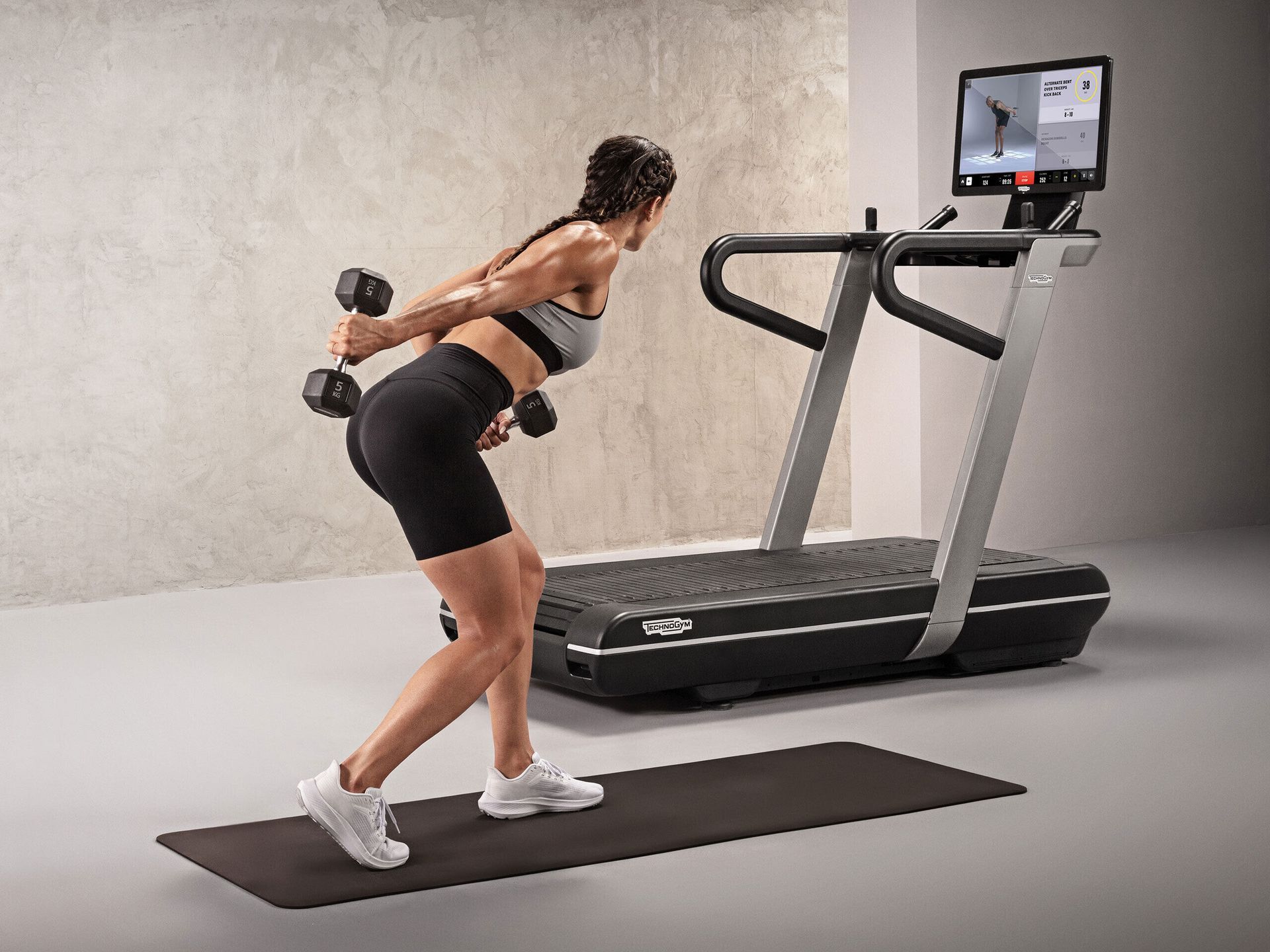 A person performs tricep kickbacks with dumbbells while standing on a mat near a Technogym treadmill in a studio.