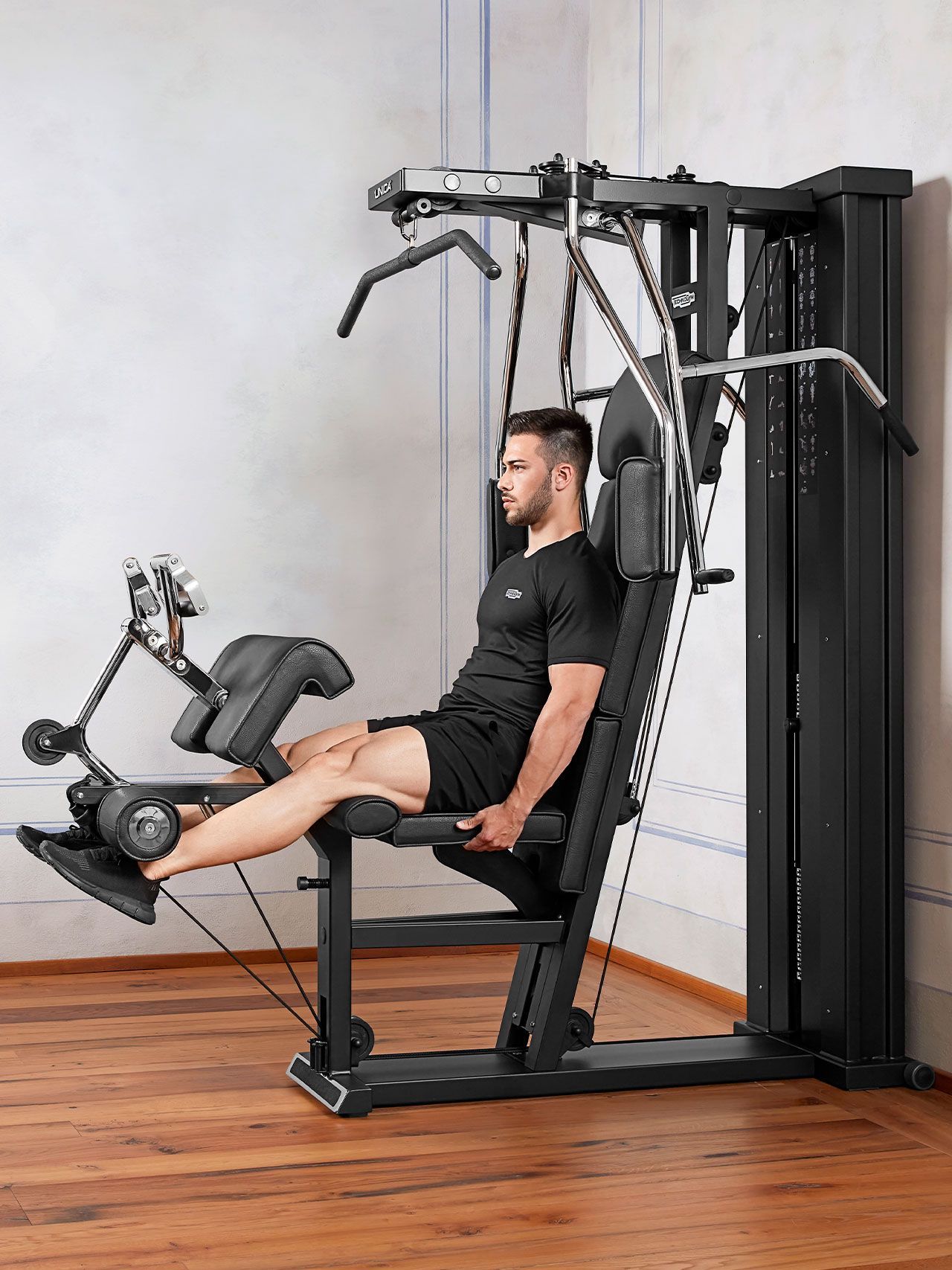 A person sitting on a black multi-gym machine, performing a leg extension exercise in a room with wood flooring.