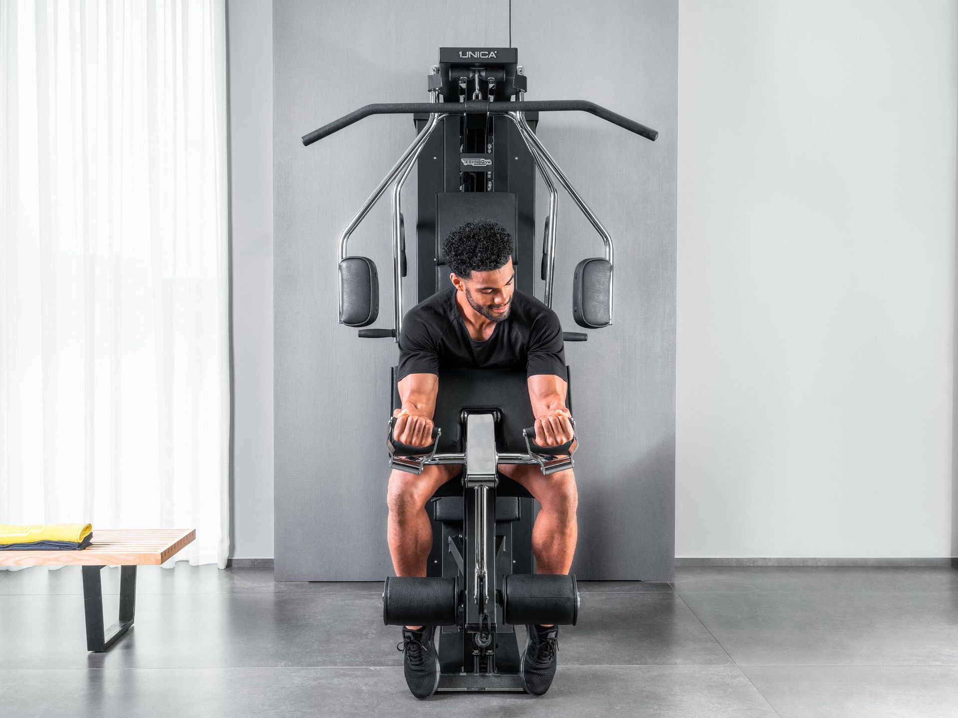 A person using a multi-station home gym machine to perform seated bicep curls in a minimalist room.