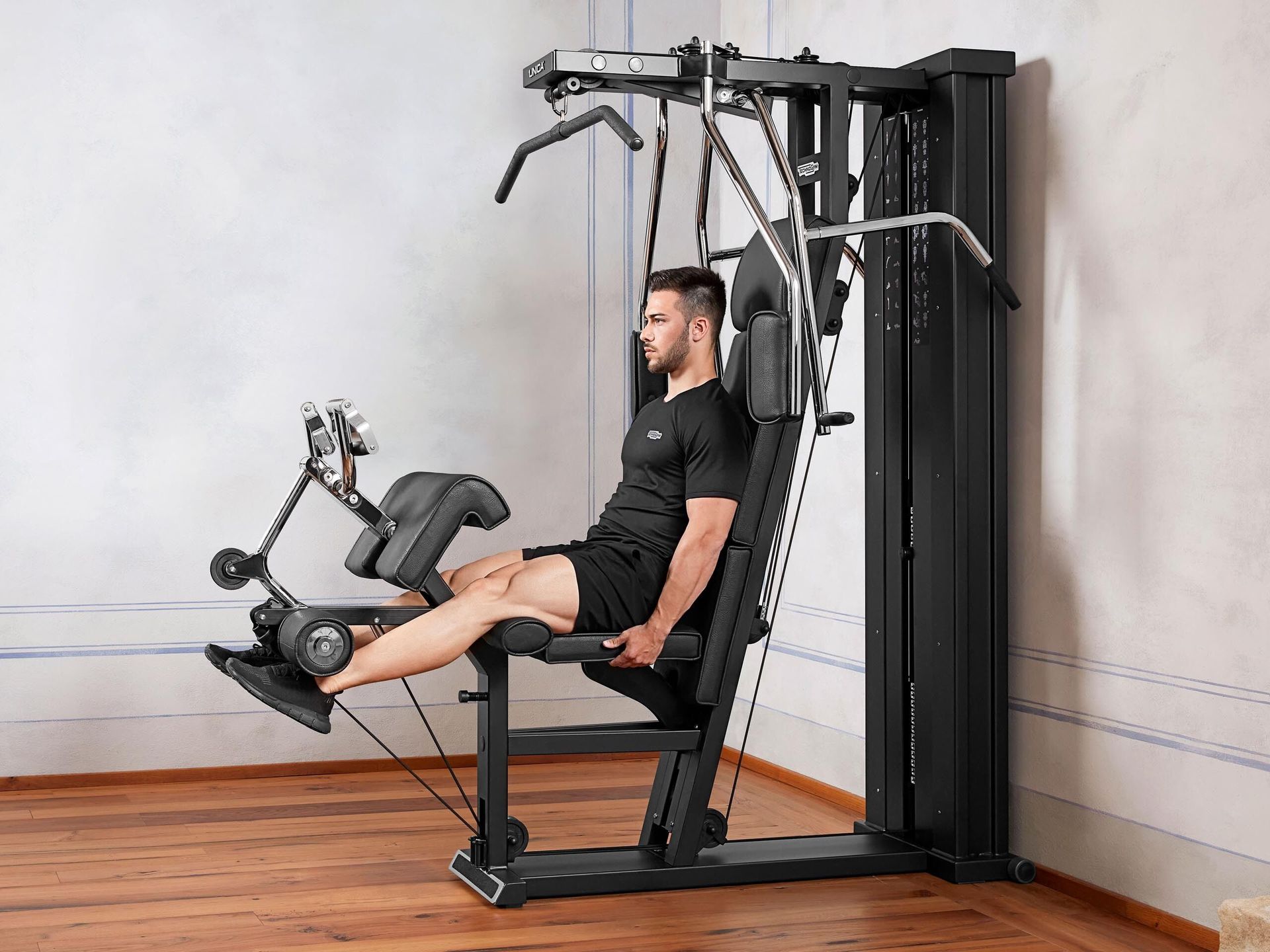 A person exercising their legs on a black multi-station home gym machine in a minimalist room with wood flooring.