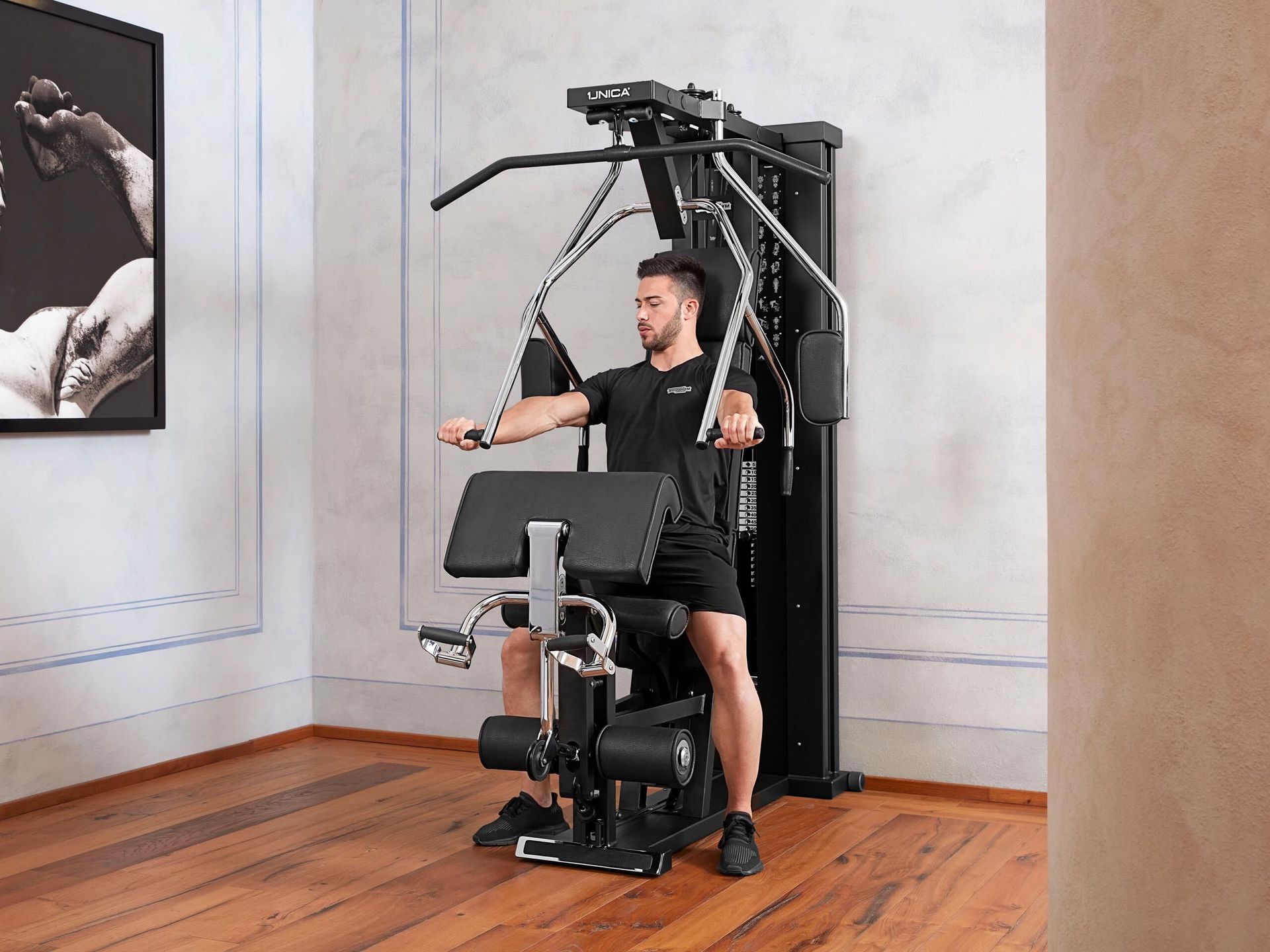 A person sitting on a black multi-station gym machine, performing a chest press exercise in a minimalist, well-lit room.