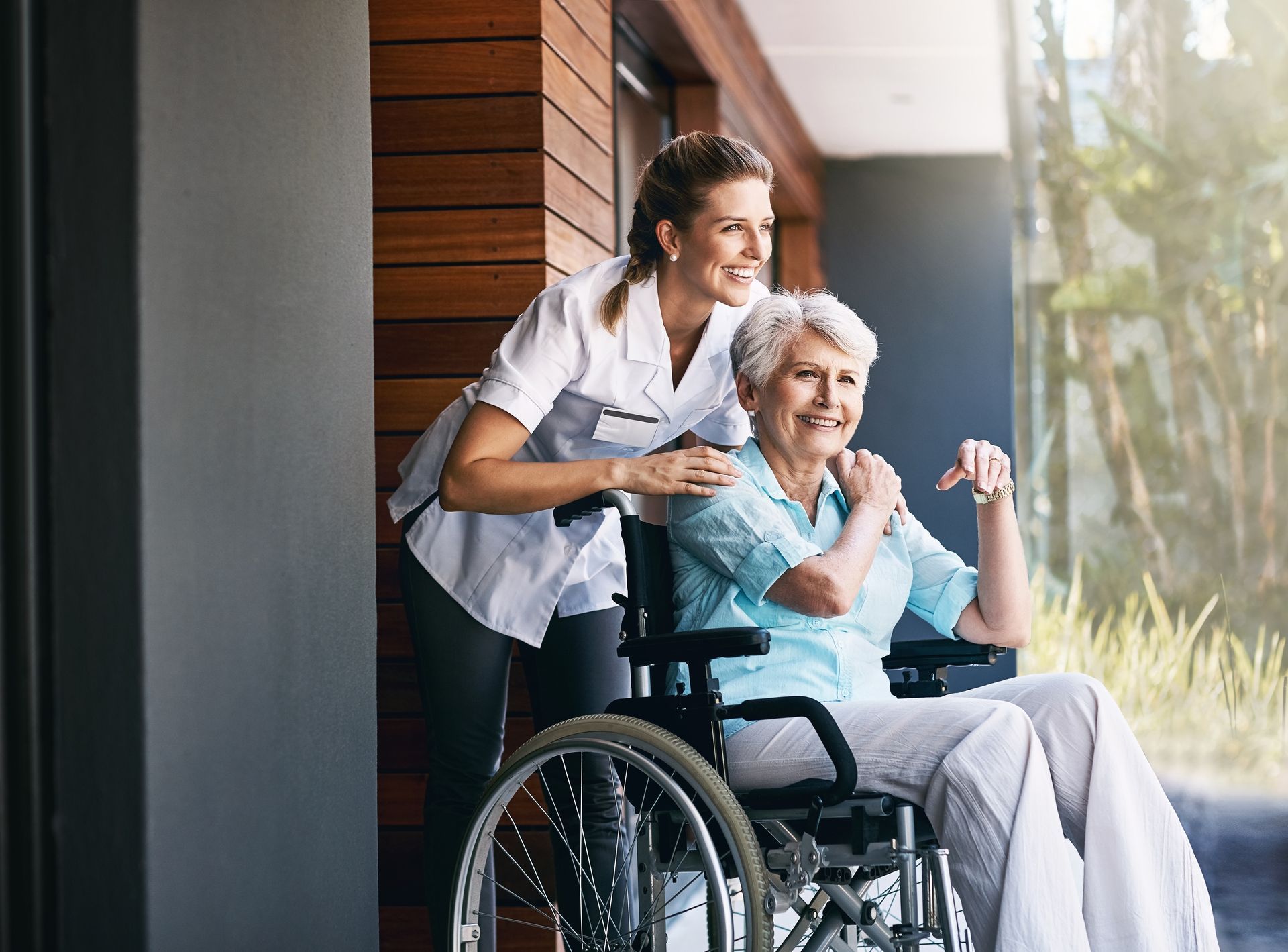Caregiver in blue scrubs helping an elderly woman stand by a window with books and a lamp nearby