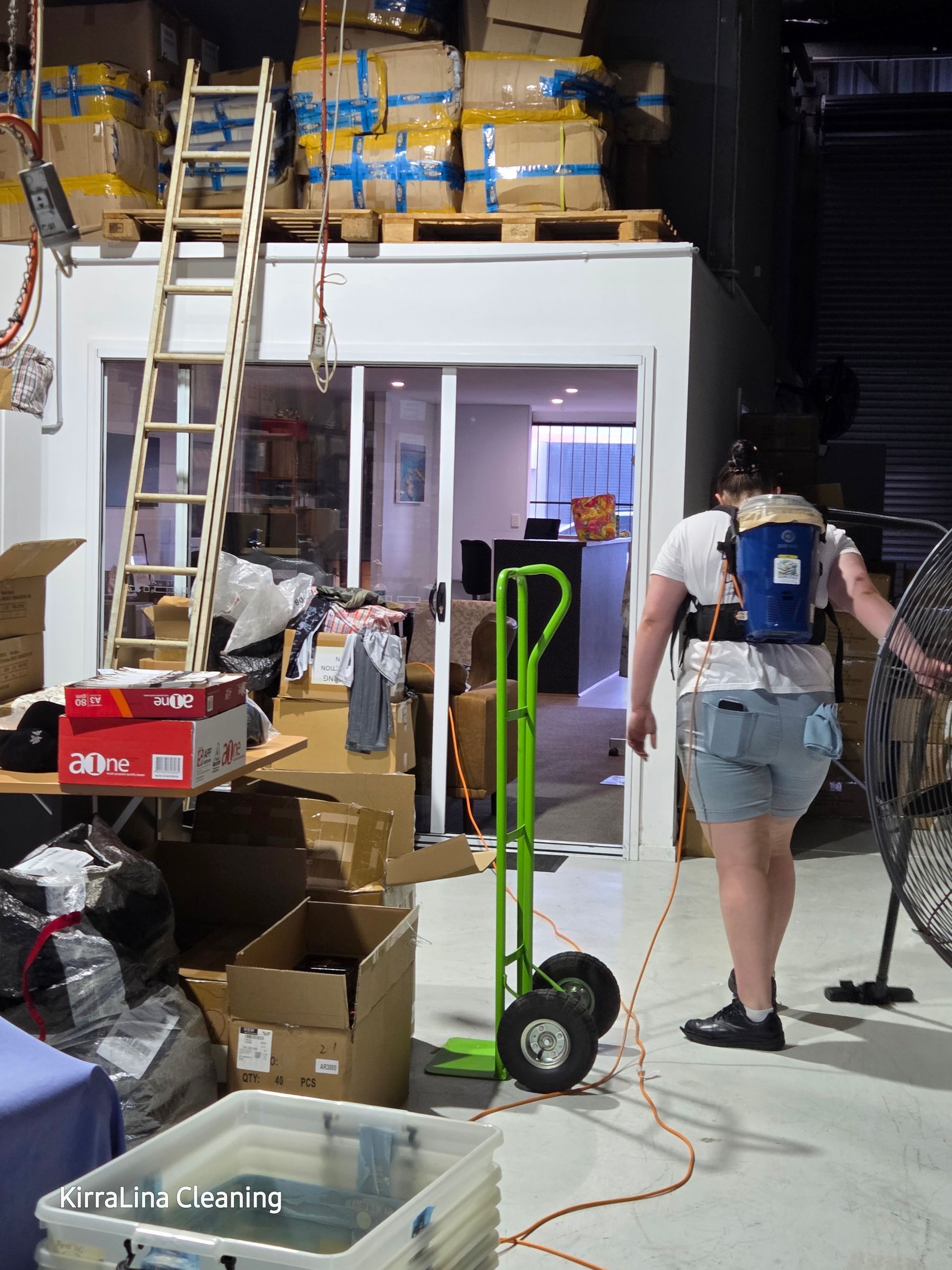 A person wearing a blue backpack vacuum cleaner cleans the floor in a dim warehouse filled with cardboard boxes — KirraLina Cleaning Services in Tweed Heads, NSW