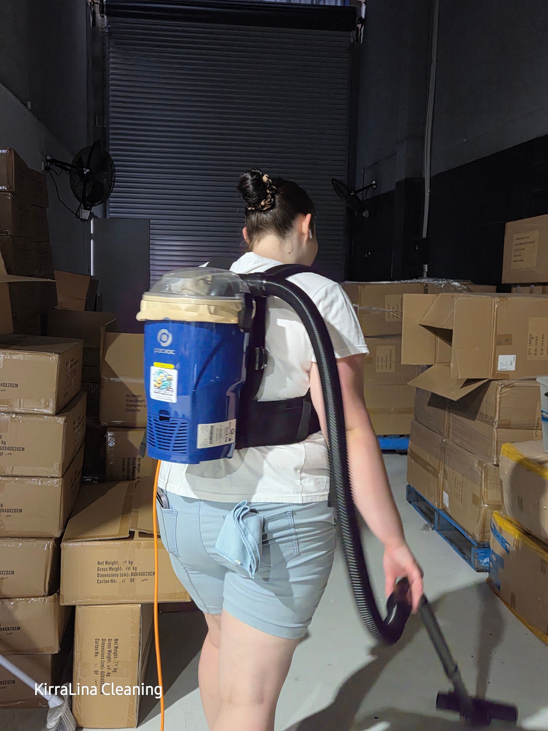 A person wearing a blue backpack vacuum cleaner cleans the floor in a dim warehouse filled with cardboard boxes — KirraLina Cleaning Services in Tweed Heads, NSW