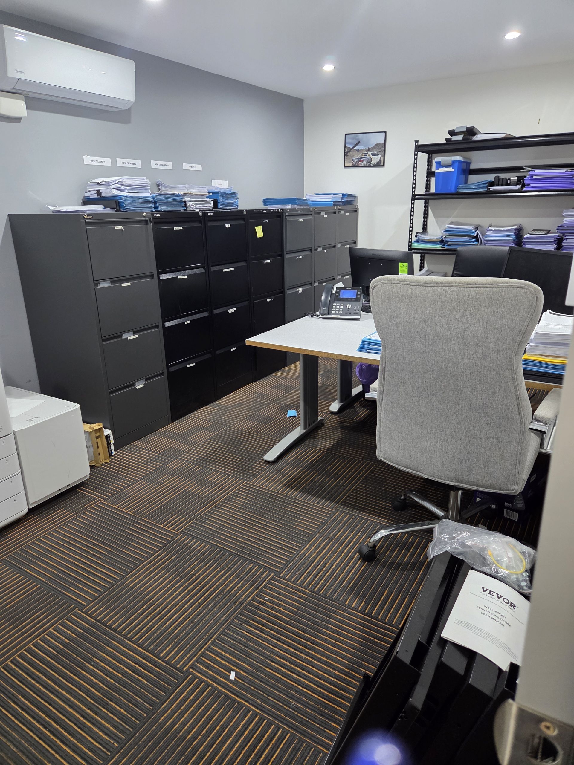 Small office with black filing cabinets, desks, grey chairs, and patterned carpet under recessed lights.