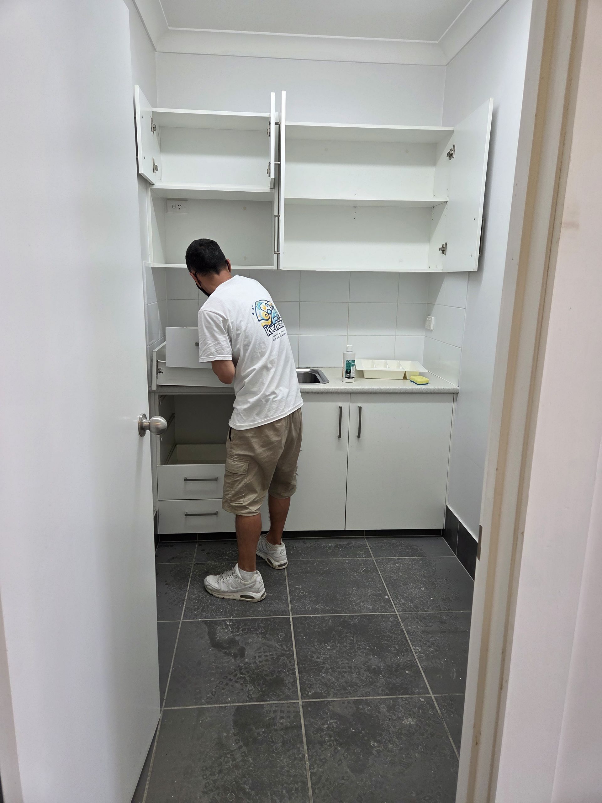 Person cleaning a small white utility room with shelves and cabinets, holding a broom.