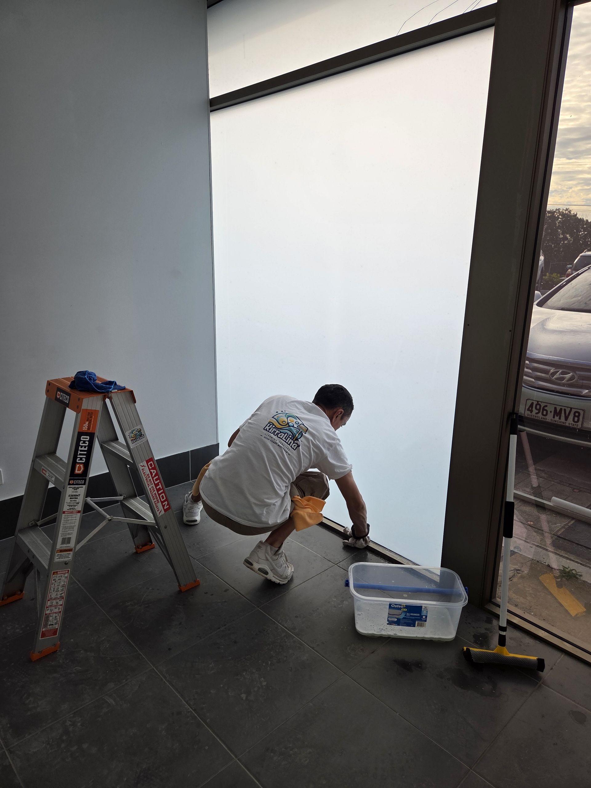 Worker kneeling beside tools near a large window, installing or repairing a dark floor inside a room.