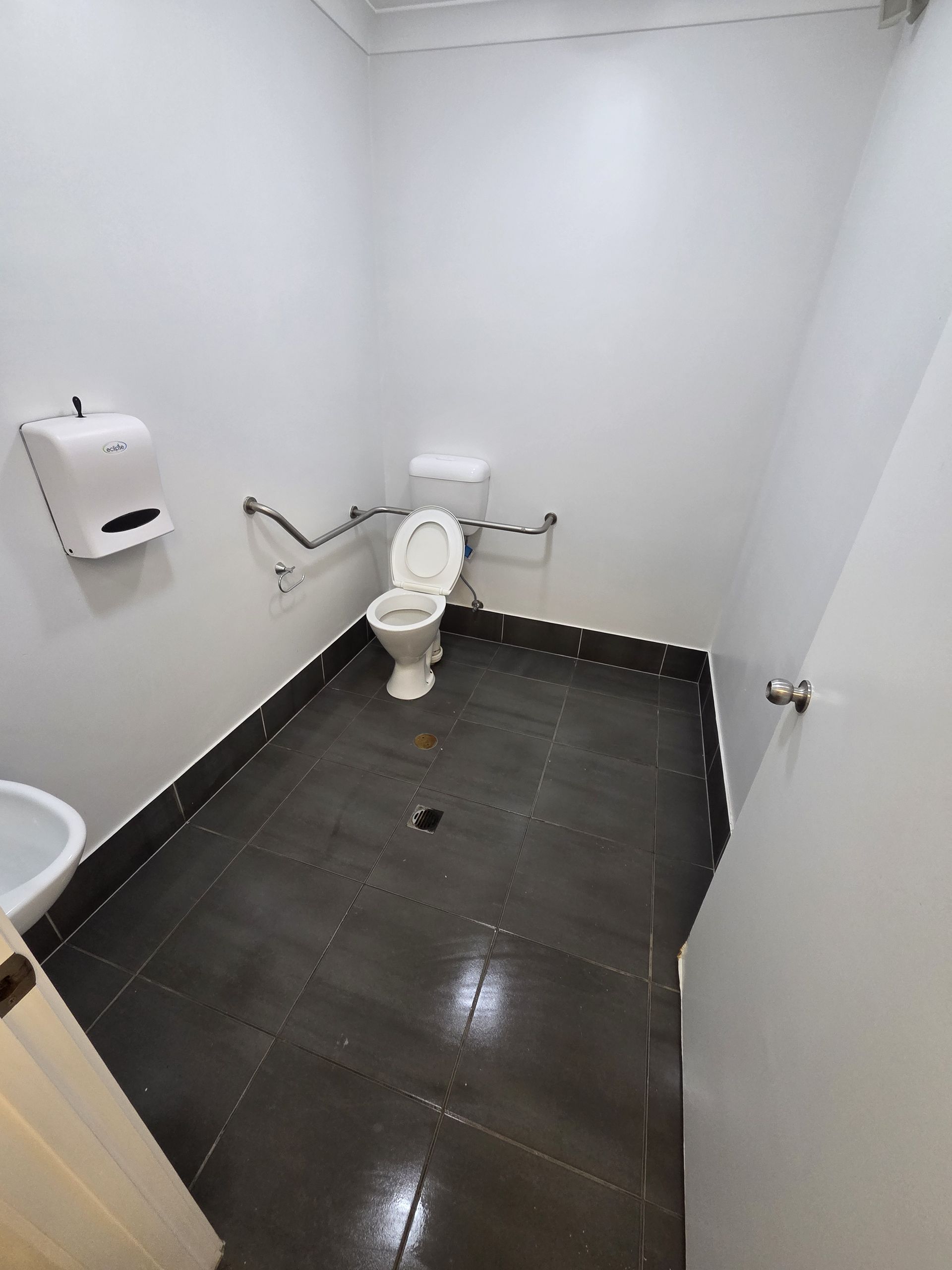 Small white bathroom with a toilet, wall dispenser, and dark tiled floor.