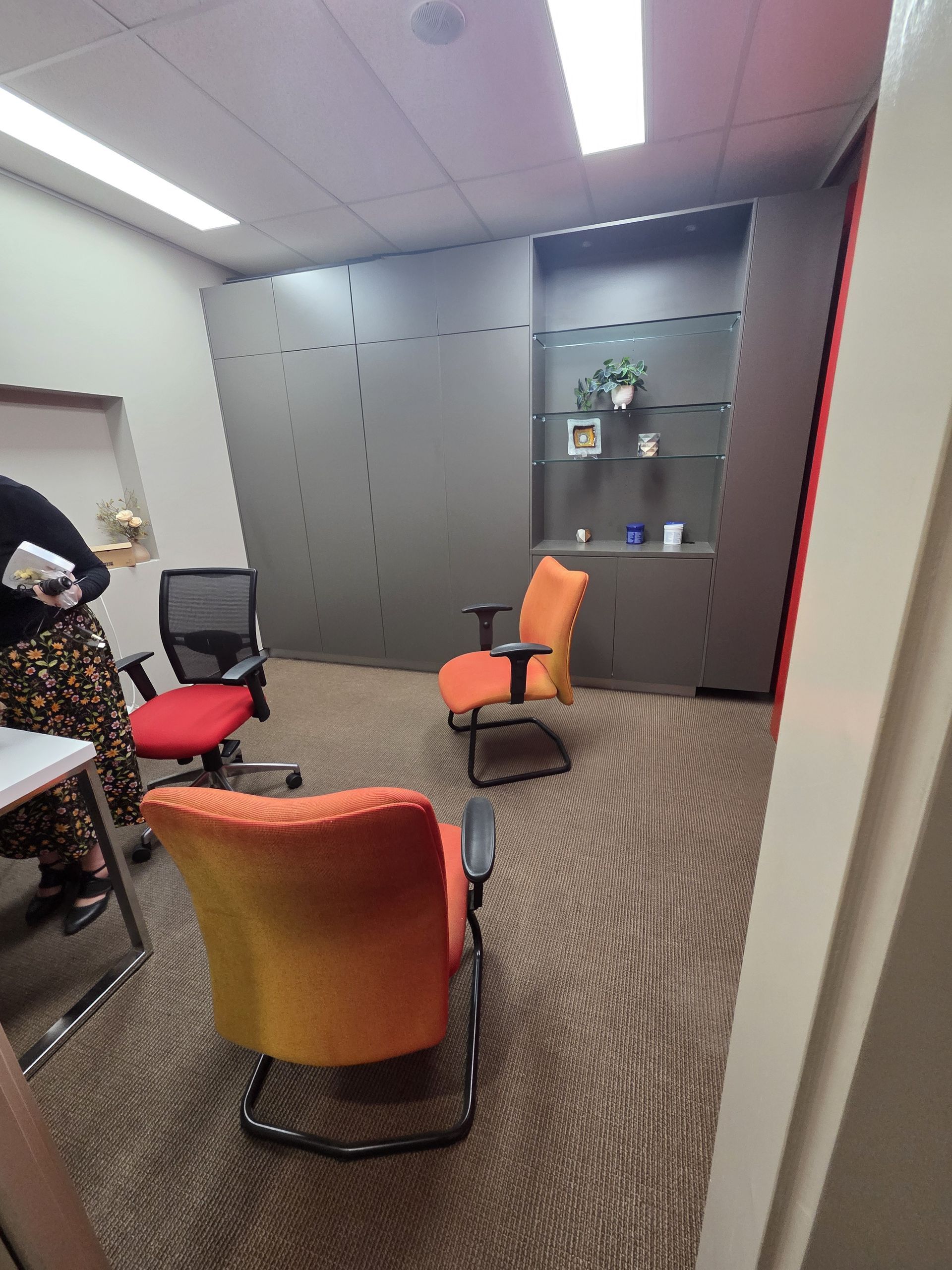 Small office with orange and red chairs, patterned carpet, cabinets, and a person at a desk on the left.