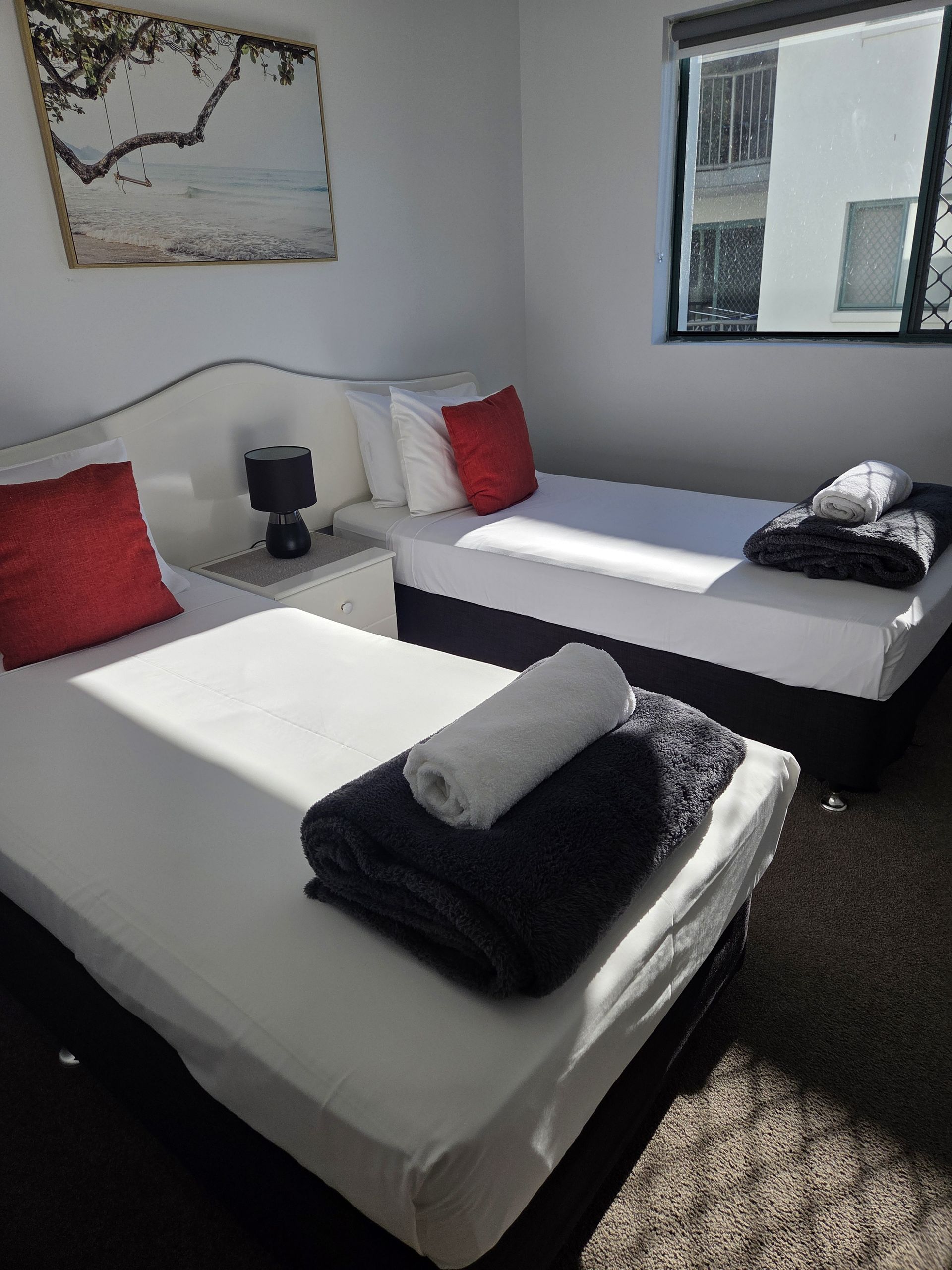 Two white beds in a bright hotel room with red pillows, black towels, and a wall mirror.