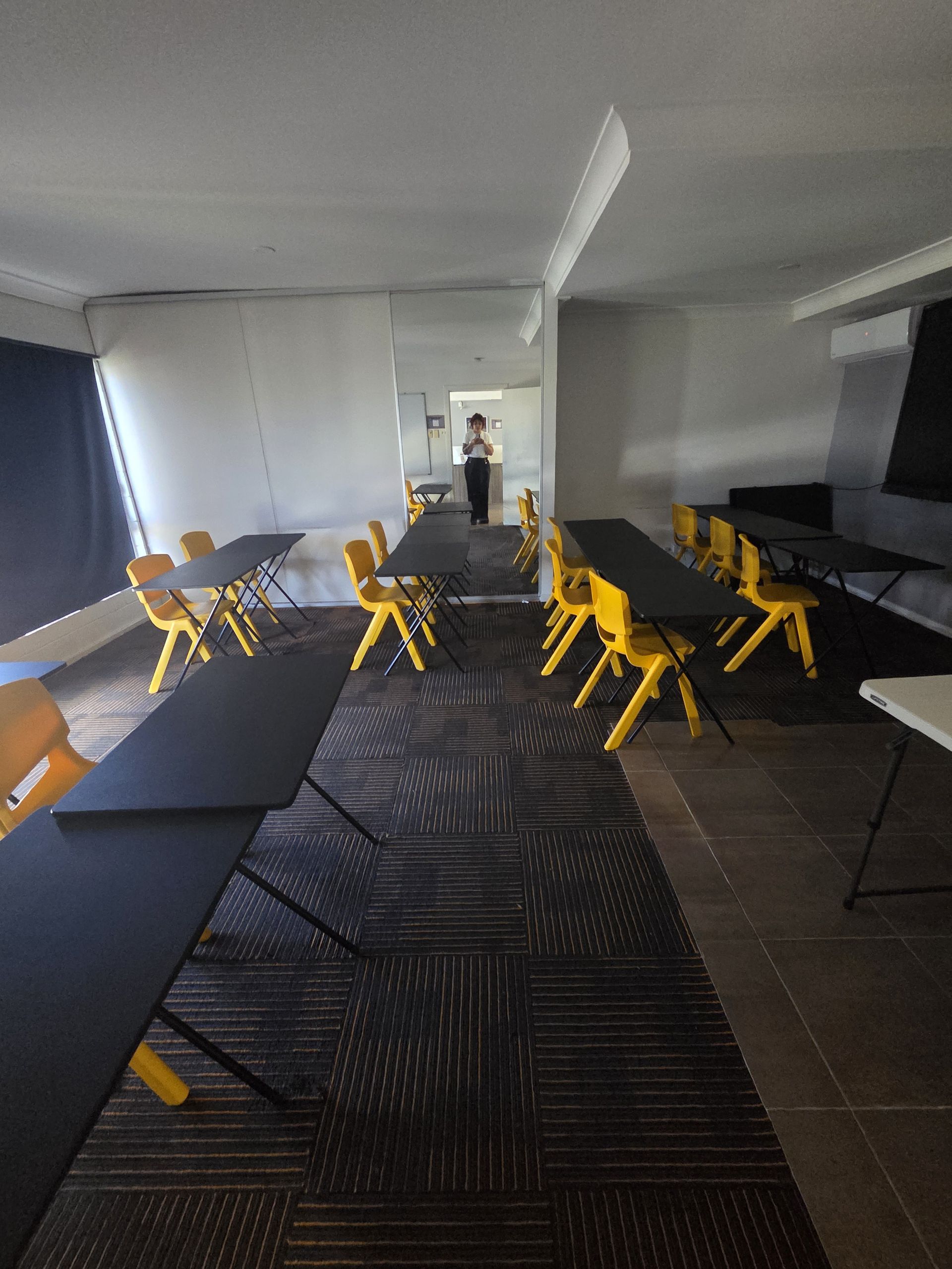 Empty meeting room with black chairs, yellow stools, and long tables on patterned carpet, viewed toward a doorway.