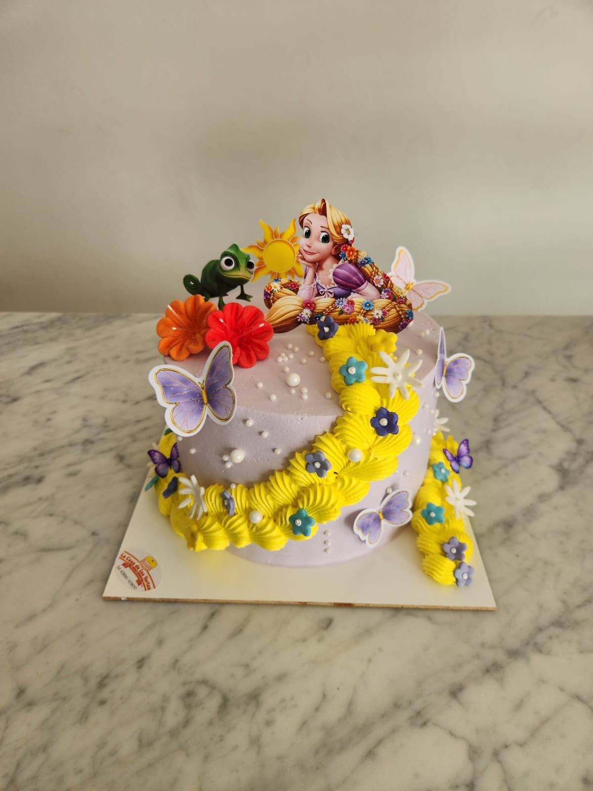 Pastel de lavanda con guirnalda floral amarilla, mariposas y figuritas coloridas sobre una mesa de mármol.