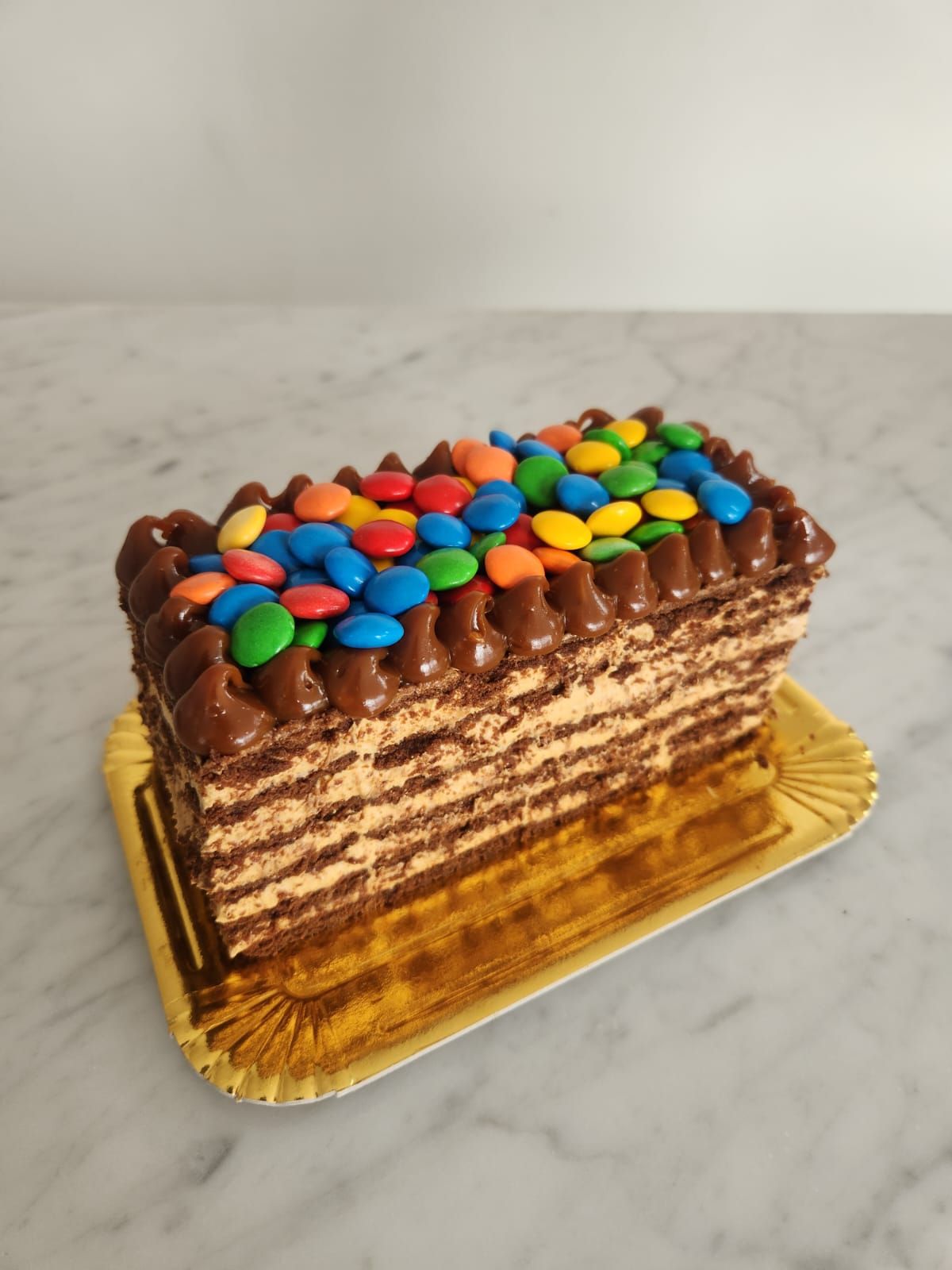 Pastel de chocolate en capas cubierto con glaseado de chocolate y trozos de caramelos de colores en una bandeja dorada.