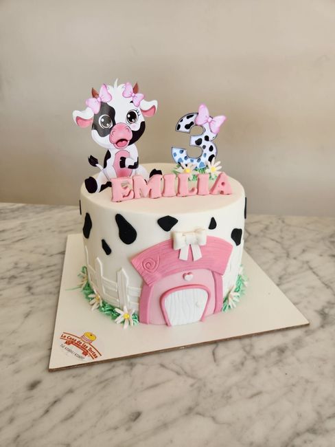 Tarta de cumpleaños con temática de vaca, con detalles en rosa y adorno con el nombre, sobre una encimera de mármol.
